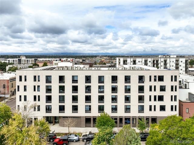 Aerial view of a modern five-story apartment building with white facade and dark windows, surrounded by trees and parked cars, with cityscape and mountains in the background under a partly cloudy sky.